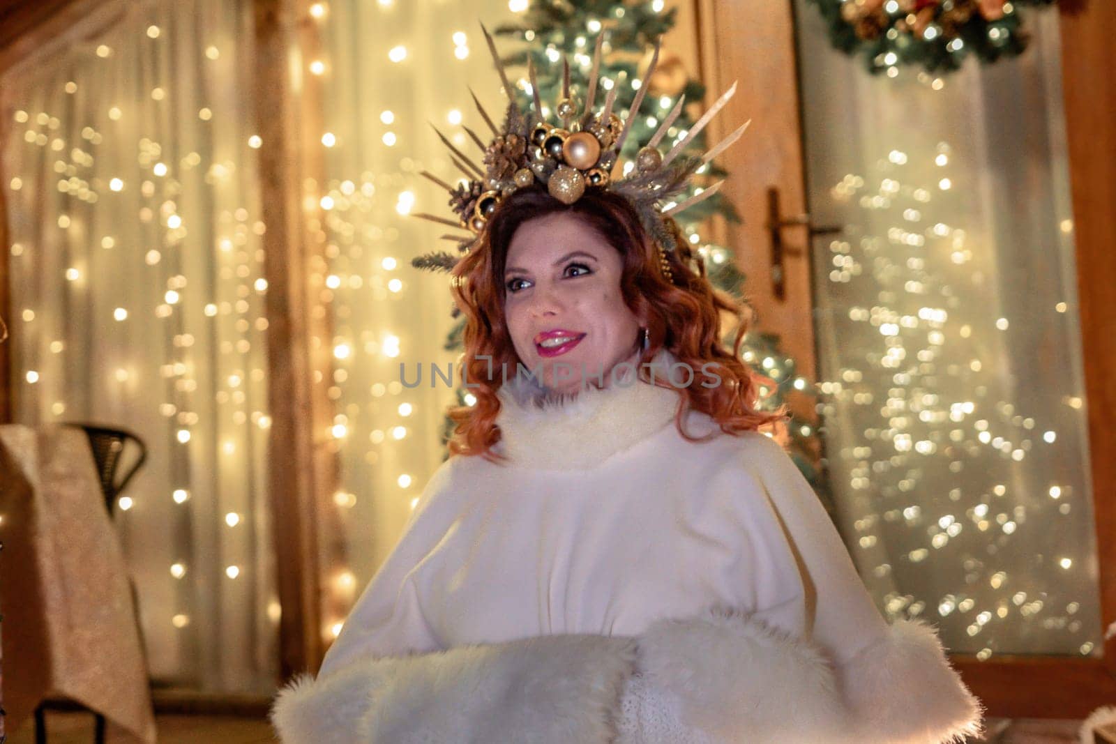 A woman with red hair wears a white fur coat and a gold crown, standing in front of a Christmas tree with lights.