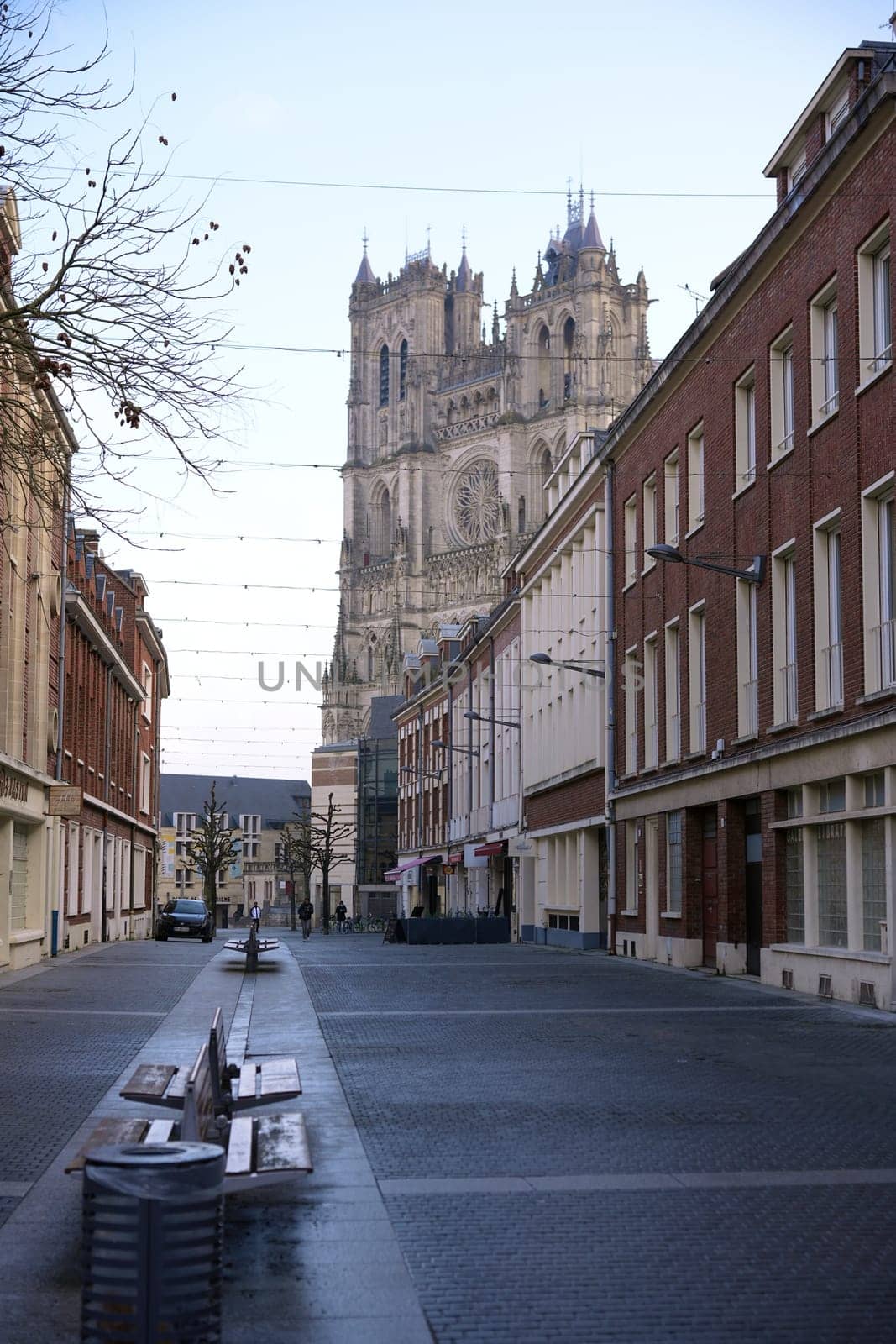 A peaceful cobblestone street lined with red brick buildings leads to a majestic Gothic cathedral under a clear sky. Amiens. France 17.02.2025