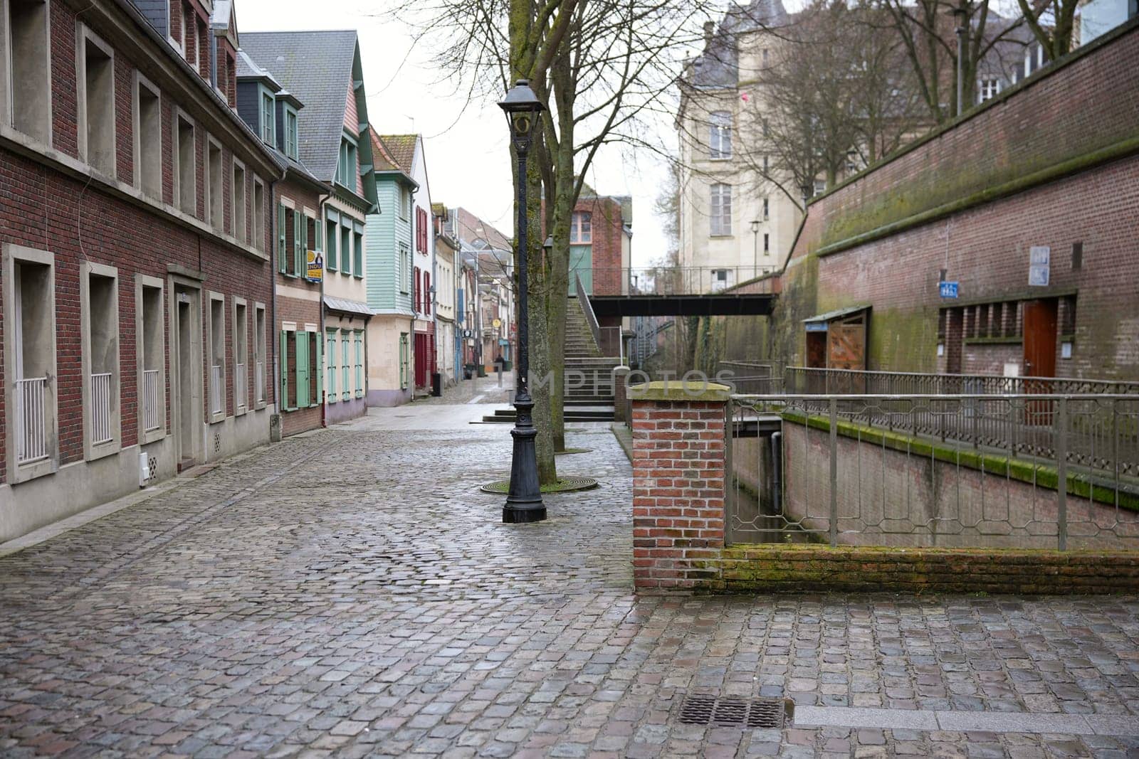 A cobblestone street in Amiens, France, lined with colorful shuttered houses, brick walls, vintage lampposts, and a serene canal, creating a historic charm