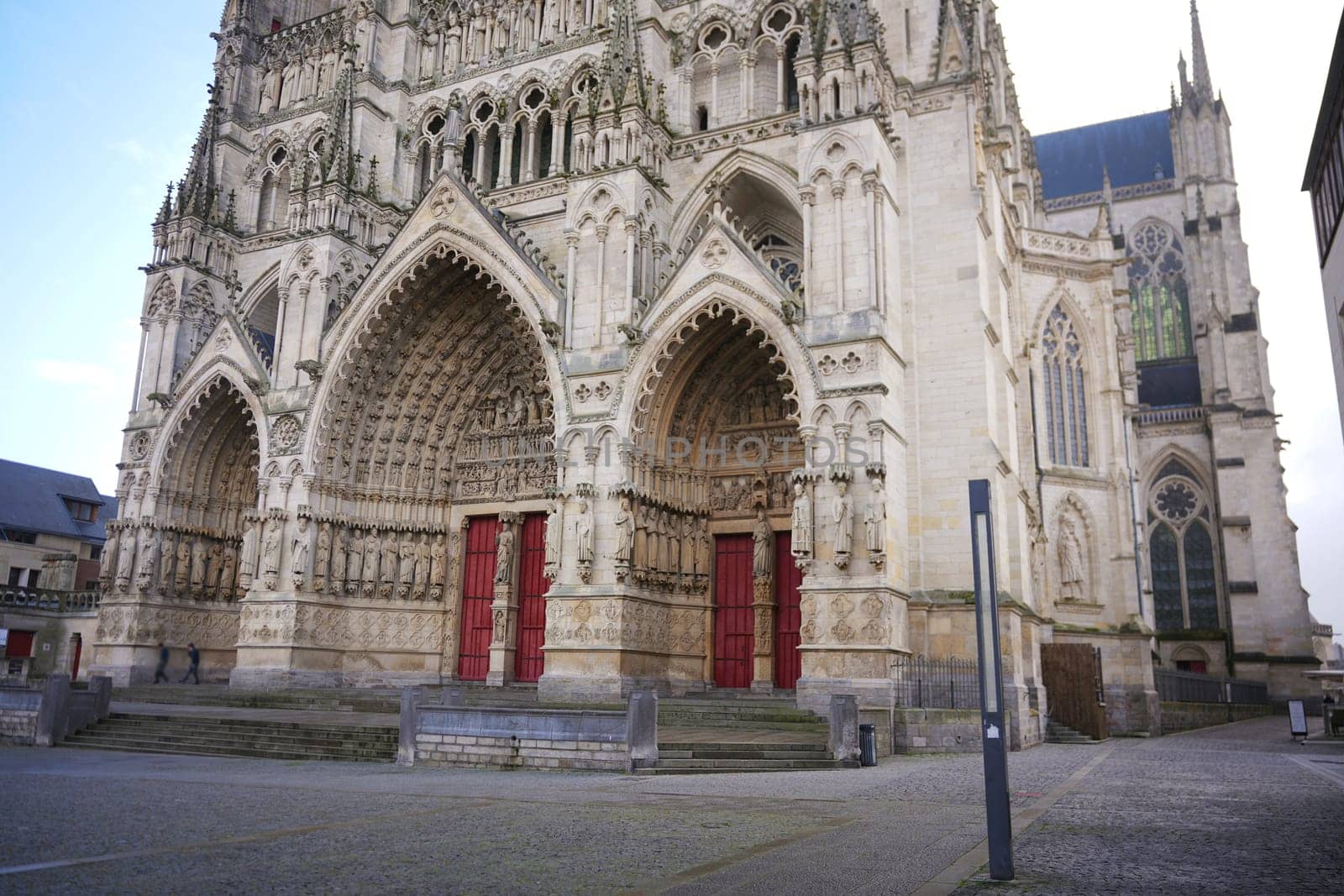 Exterior view of a Gothic cathedral highlighting ornate arches, intricate sculptures, and vibrant red entry doors on a clear day. Amiens. France 17.02.2025