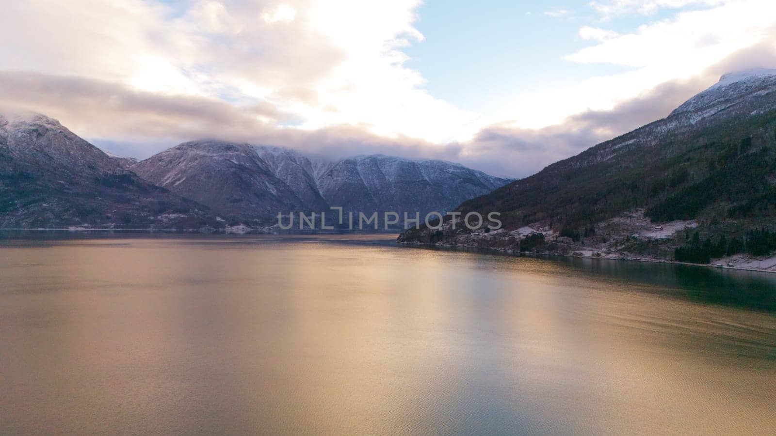 Aerial winter view of Gaupne, Norway, featuring a fjord reflecting golden sunset light, surrounded by snow-covered mountains and a peaceful rural landscape.