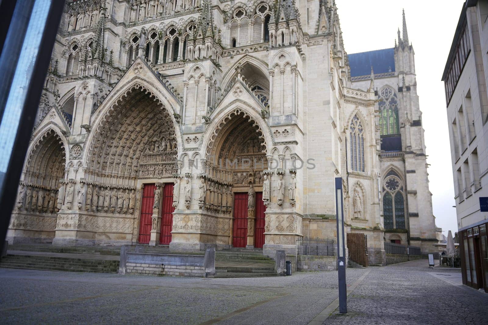 Wide view of a Gothic cathedral facade featuring intricately carved portals, red doors, and towering architectural details. Amiens. France 17.02.2025