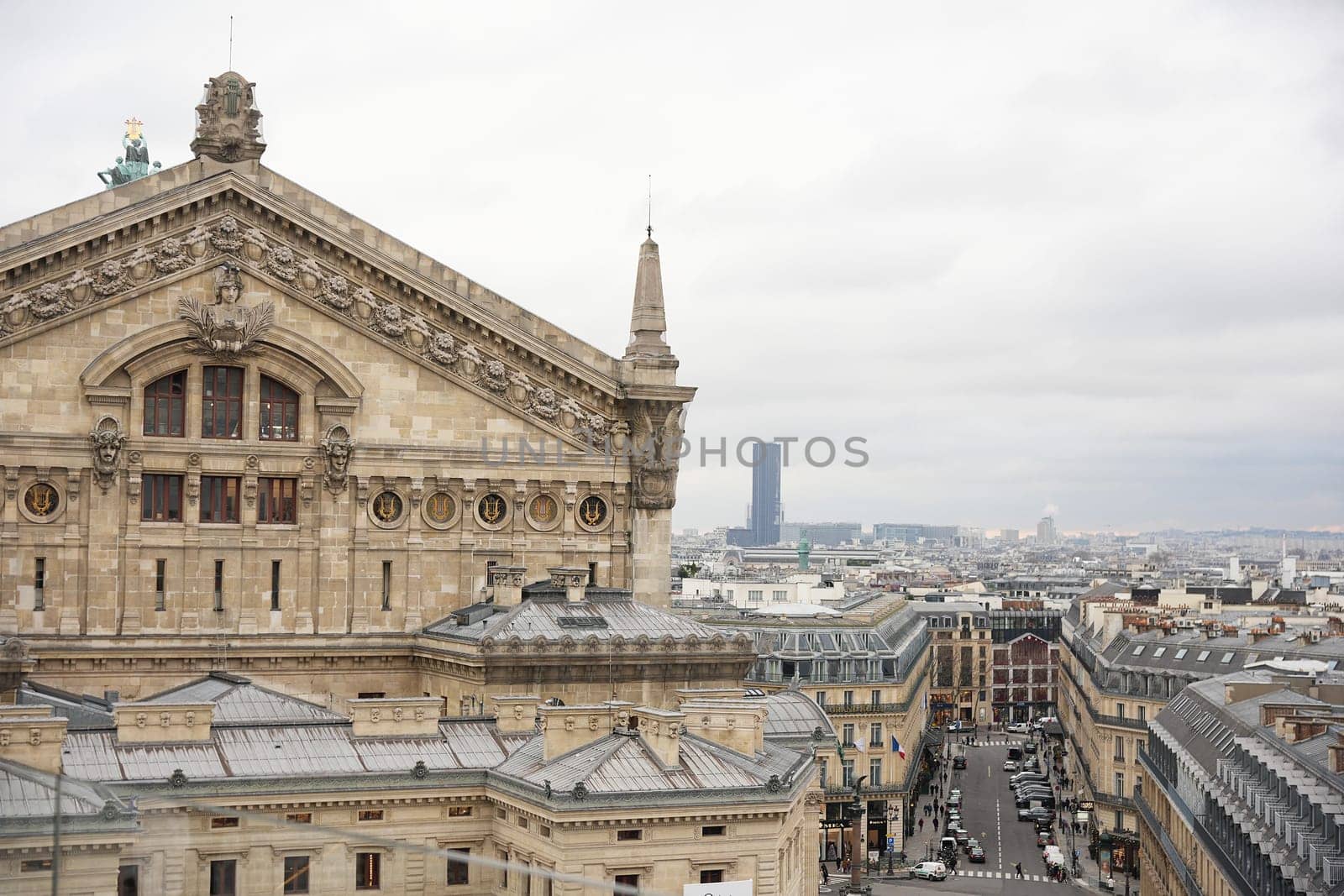 A close-up of the intricate facade of the Opera Garnier in Paris, with classic rooftops, bustling streets, and a modern skyscraper in the background under cloudy skies.