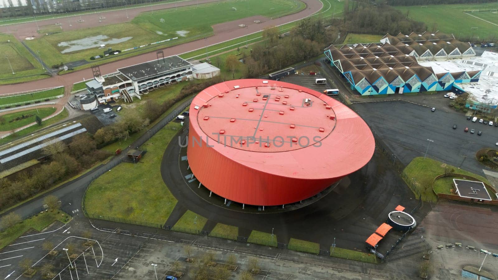 Aerial view of a striking red circular building with a unique design, surrounded by green fields, parking, and nearby facilities. A modern architectural landmark.