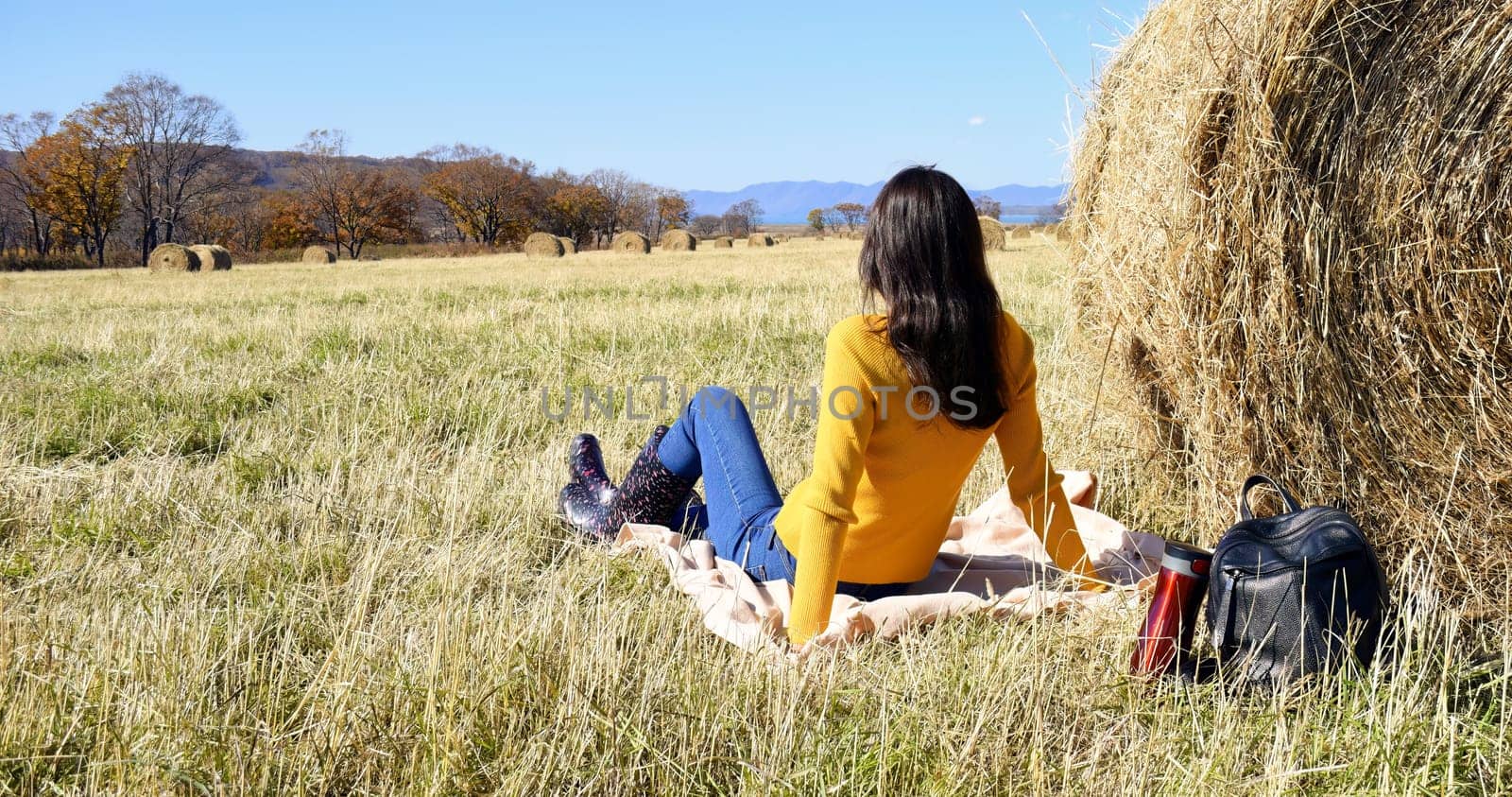 Woman in yellow sweater and rubber boots sitting on plaid next to backpack and thermos with hot tea near haystack in field. she looking thoughtfully into the distance mountains and the autumn forest.