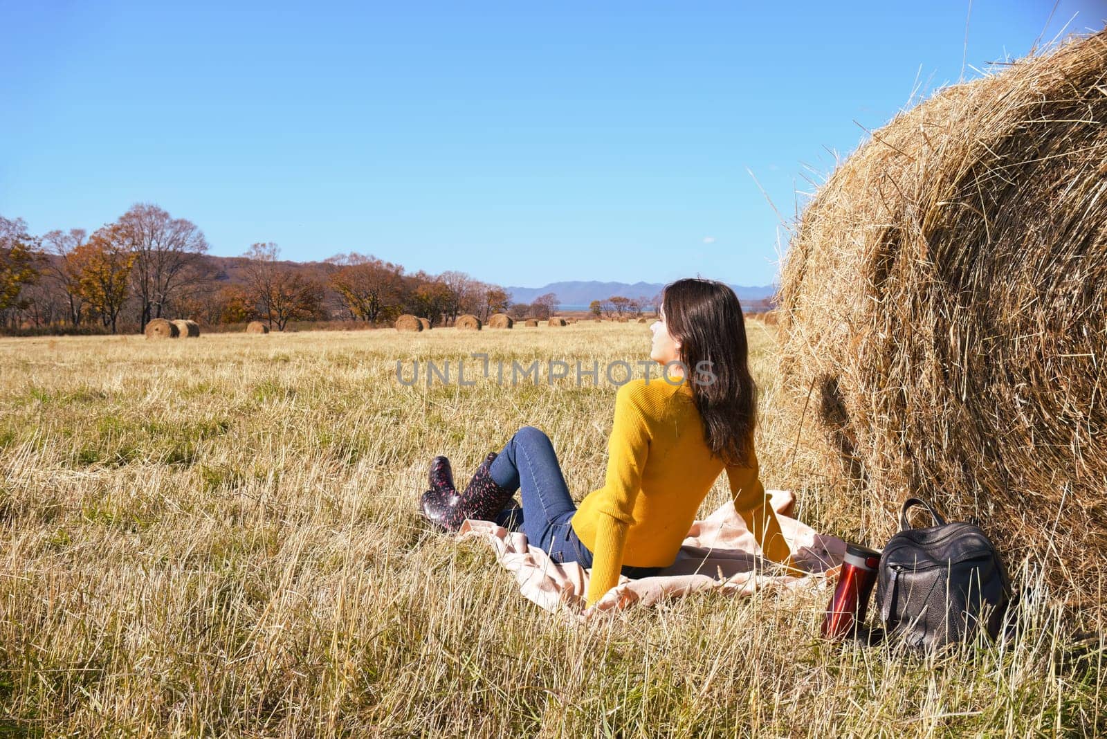 Woman in yellow sweater and rubber boots sitting on plaid next to backpack and thermos with hot tea near haystack in field. she looking thoughtfully into the distance mountains and the autumn forest.