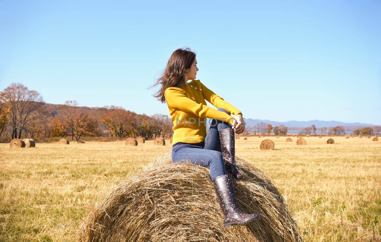 Young brunette woman in yellow sweater and rubber boots sitting on top of haystack in field. she looking at mountains, Japanese sea and the autumn forest. Primorsky kray, Russia. Beauty in nature.