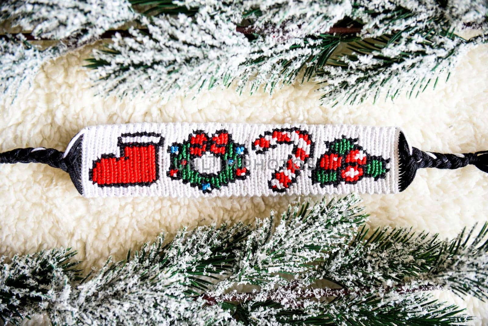 Woven friendship bracelet with pattern Christmas berry, lollipop, wreath and santa boot. Handmade of thread, on  background with snowy fir branches. DIY New Year gift idea
