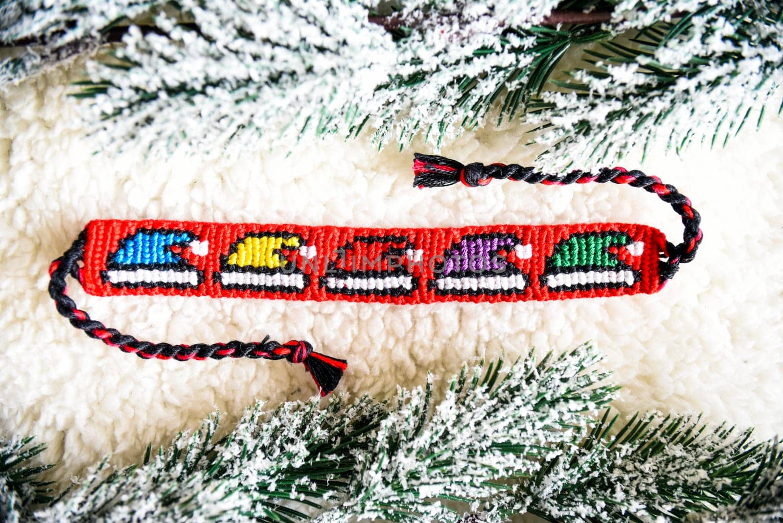 Woven friendship bracelet with bright alpha pattern Multicolored christmas hats, handmade of thread, next to snowy fir branches.