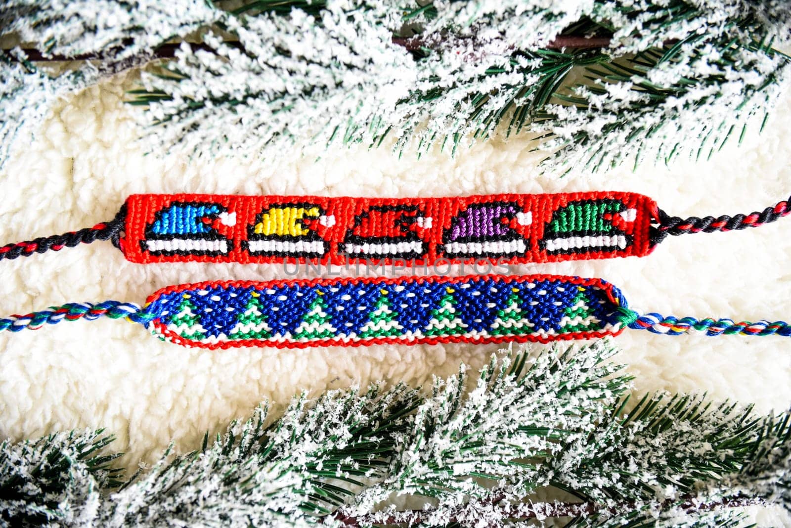 Woven friendship bracelets with alpha patterns Snowy fir trees, Santa hats. Handmade of thread on light background with  snowy branches. DIY New Year gift idea
