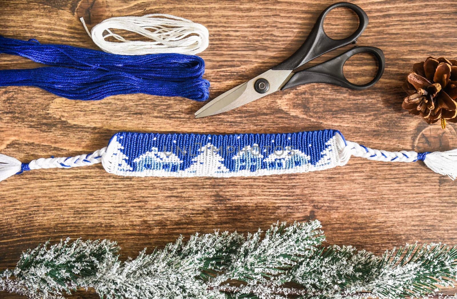 Woven friendship bracelet with alpha pattern Snowy night with houses. Handmade of thread, on wooden background with scissors, skeins of thread, spruce cone, snowy branches. DIY New Year gift idea