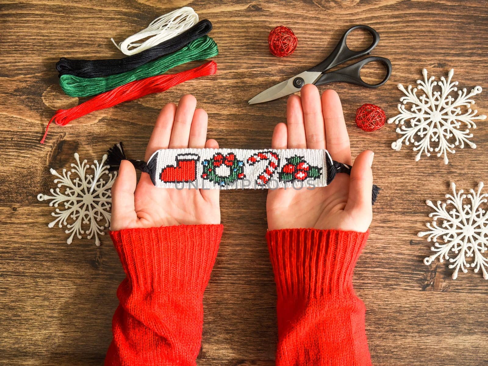 Idea for Christmas gift. Friendship bracelet with New Year alpha pattern handmade of floss threads in female hands in red sweater. Scissors, skeins of embroidery thread on wooden background.