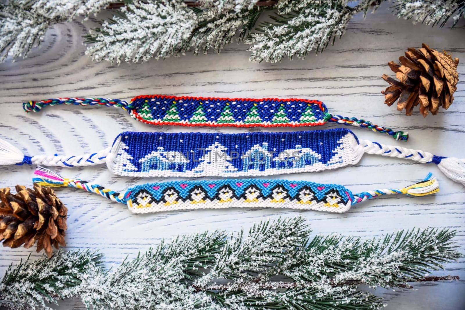Woven friendship bracelets with alpha patterns Snowy night with houses,penguins, fir trees. Handmade of thread, on wooden background with spruce cones, snowy branches.