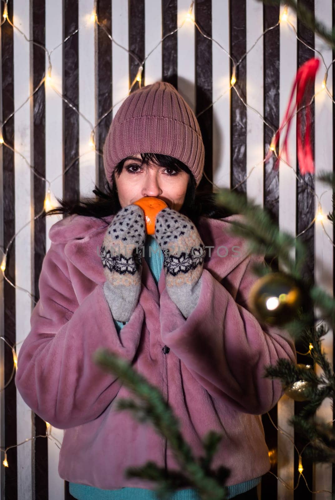Russian 48 years old woman in pink coat holding tangerines against yellow christmas lights before Christmas