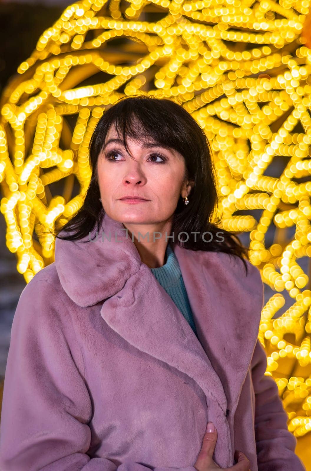 Russian 48 years old woman in blue sweater and brown coat standing against yellow christmas lights before Christmas