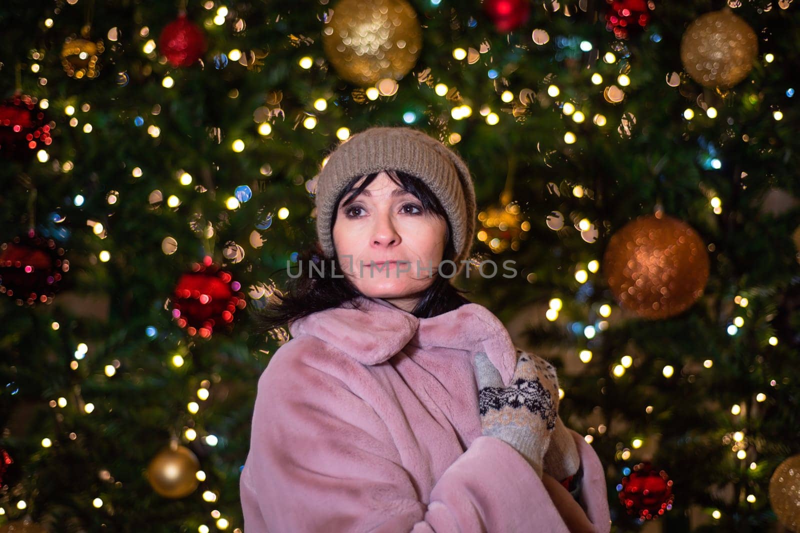 Russian 48 years old woman in pink coat standing against christmas tree and lights before New Year