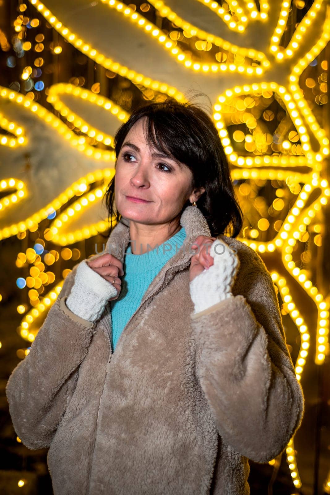 Russian 48 years old woman in blue sweater and brown coat standing against yellow christmas lights before Christmas