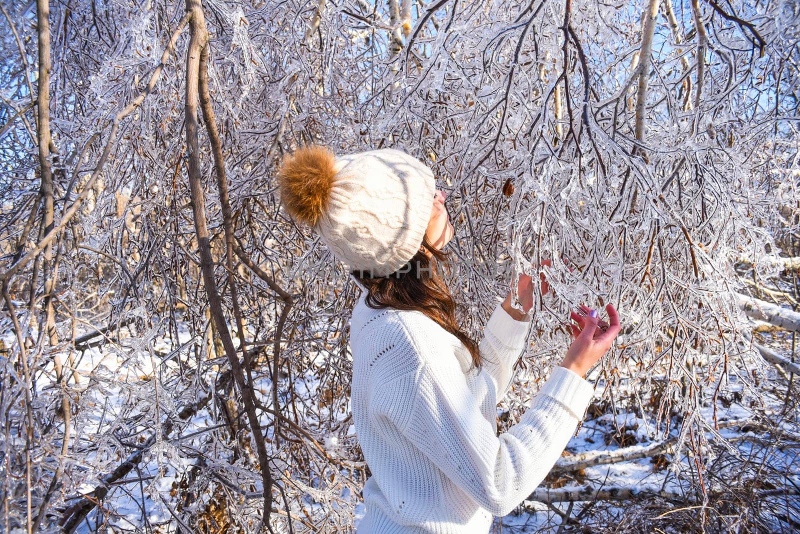 Portrait of young Russian woman in white sweater and beanie next to frozen branches of birch trees, covered with ice. Winter forest in Russia