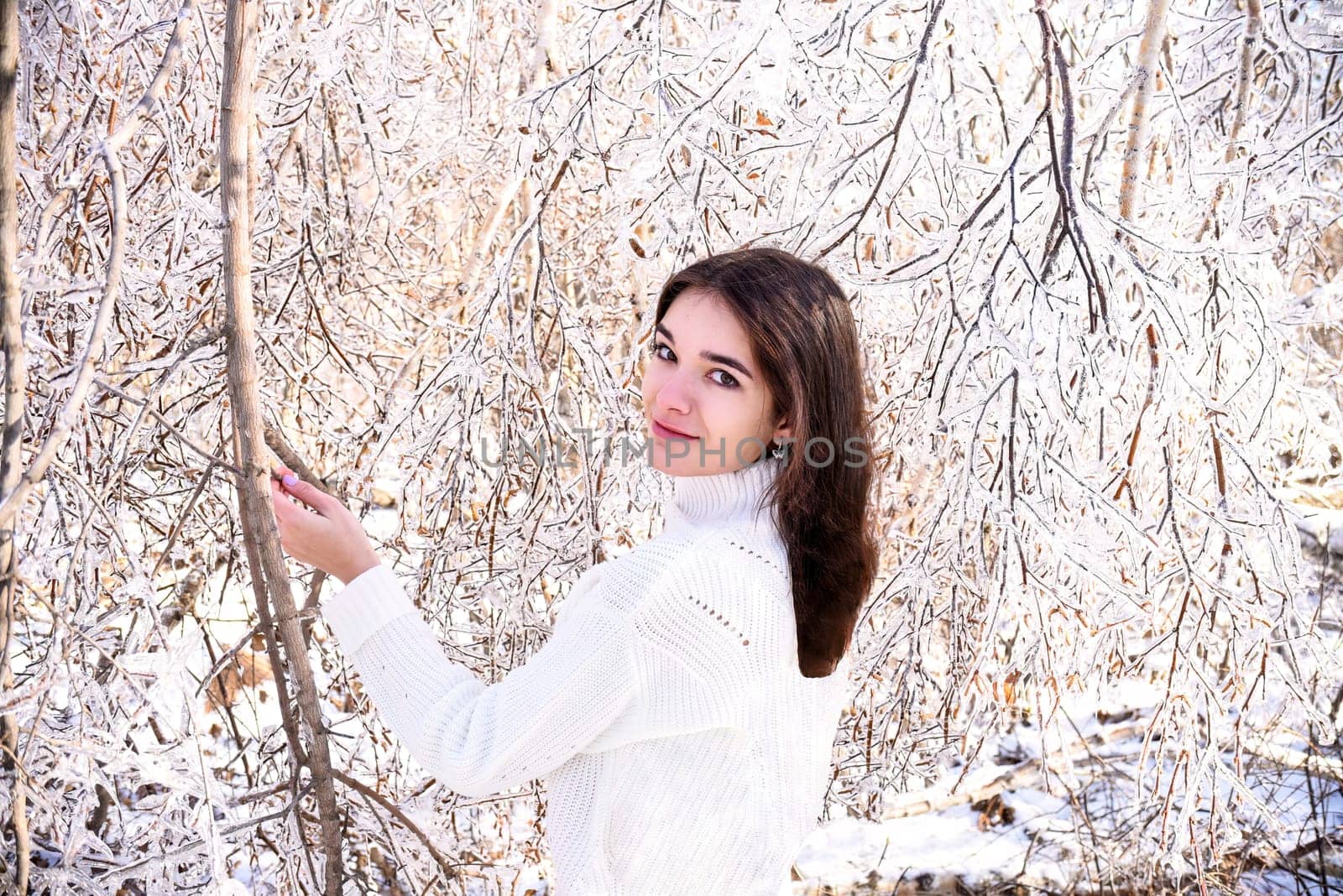 Portrait of young Russian woman in white sweater looking at camera next to frozen branches of birch trees, covered with ice. Winter forest in Russia