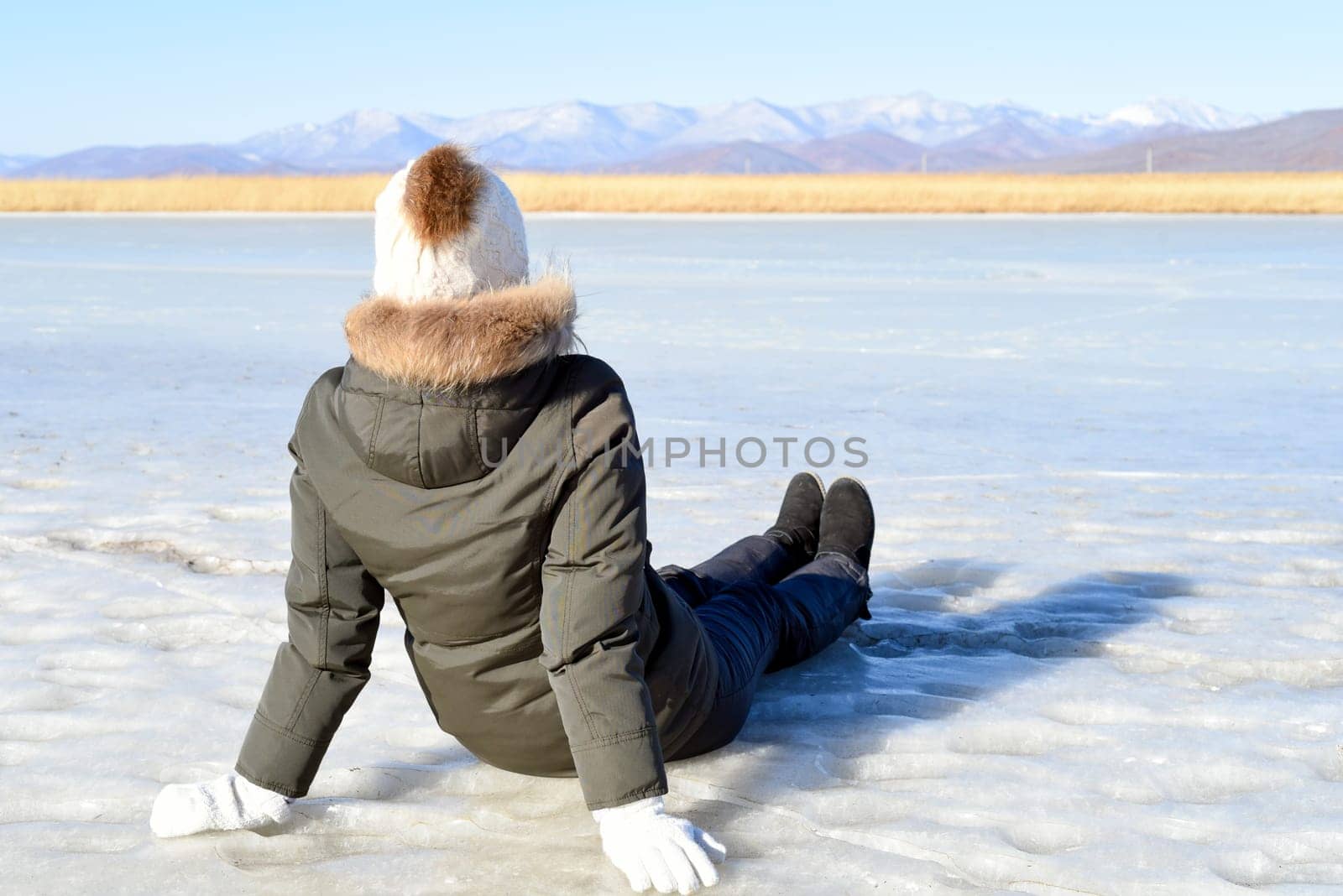 Young woman in warm clothes sitting on the ice and looking at snowy mountains. winter, Russia