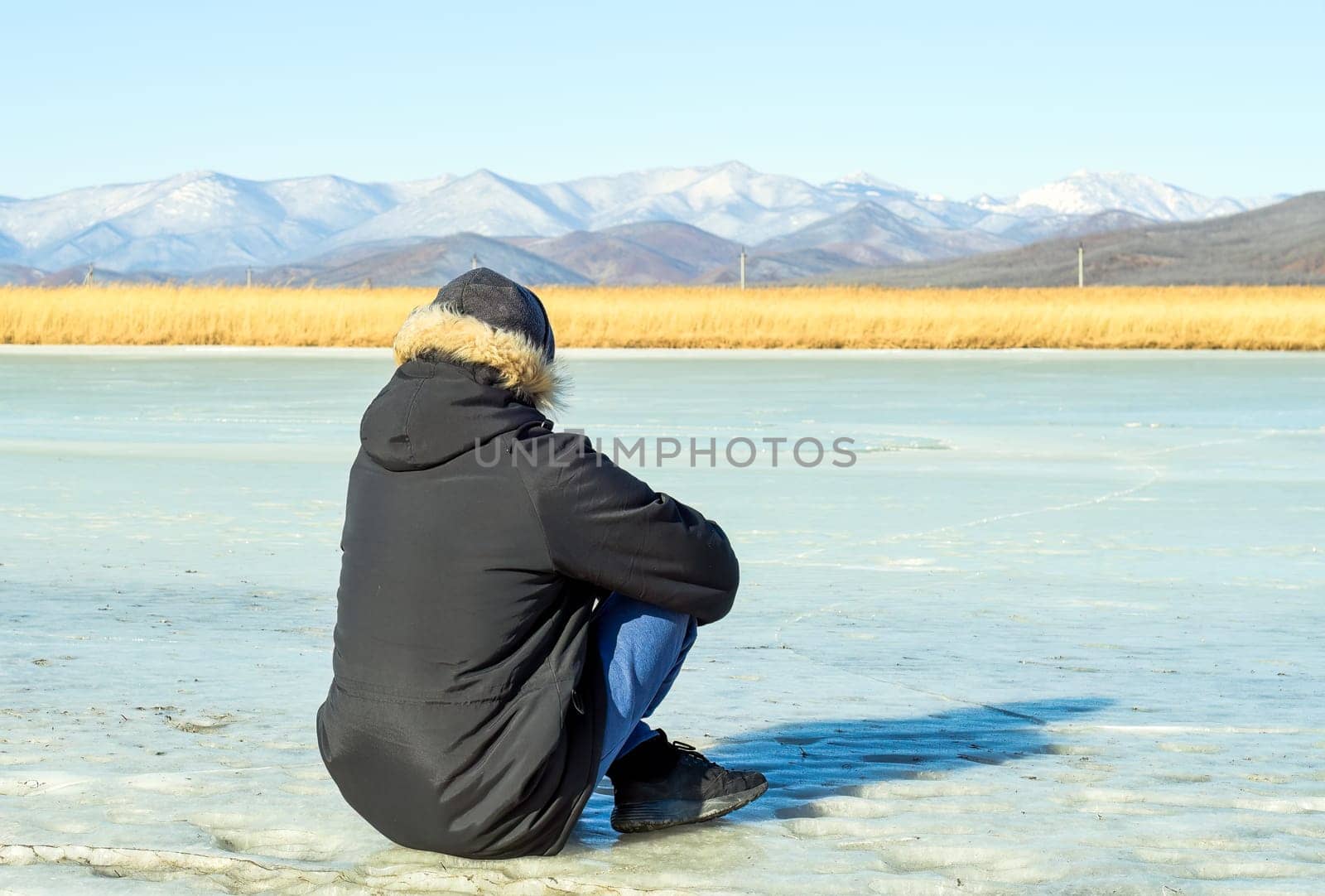 Young man in warm clothes sitting on the ice and looking at snowy mountains. winter, Russia.