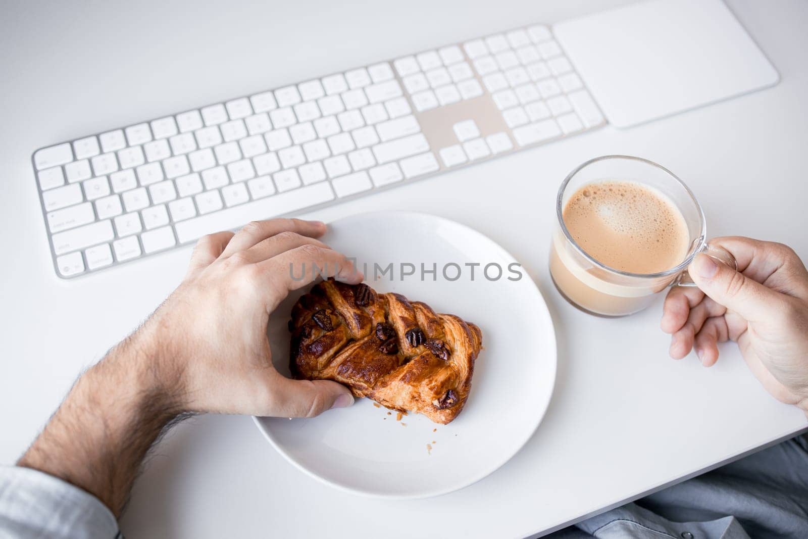Caucasian male adult enjoying coffee and pastry at office desk with keyboard.