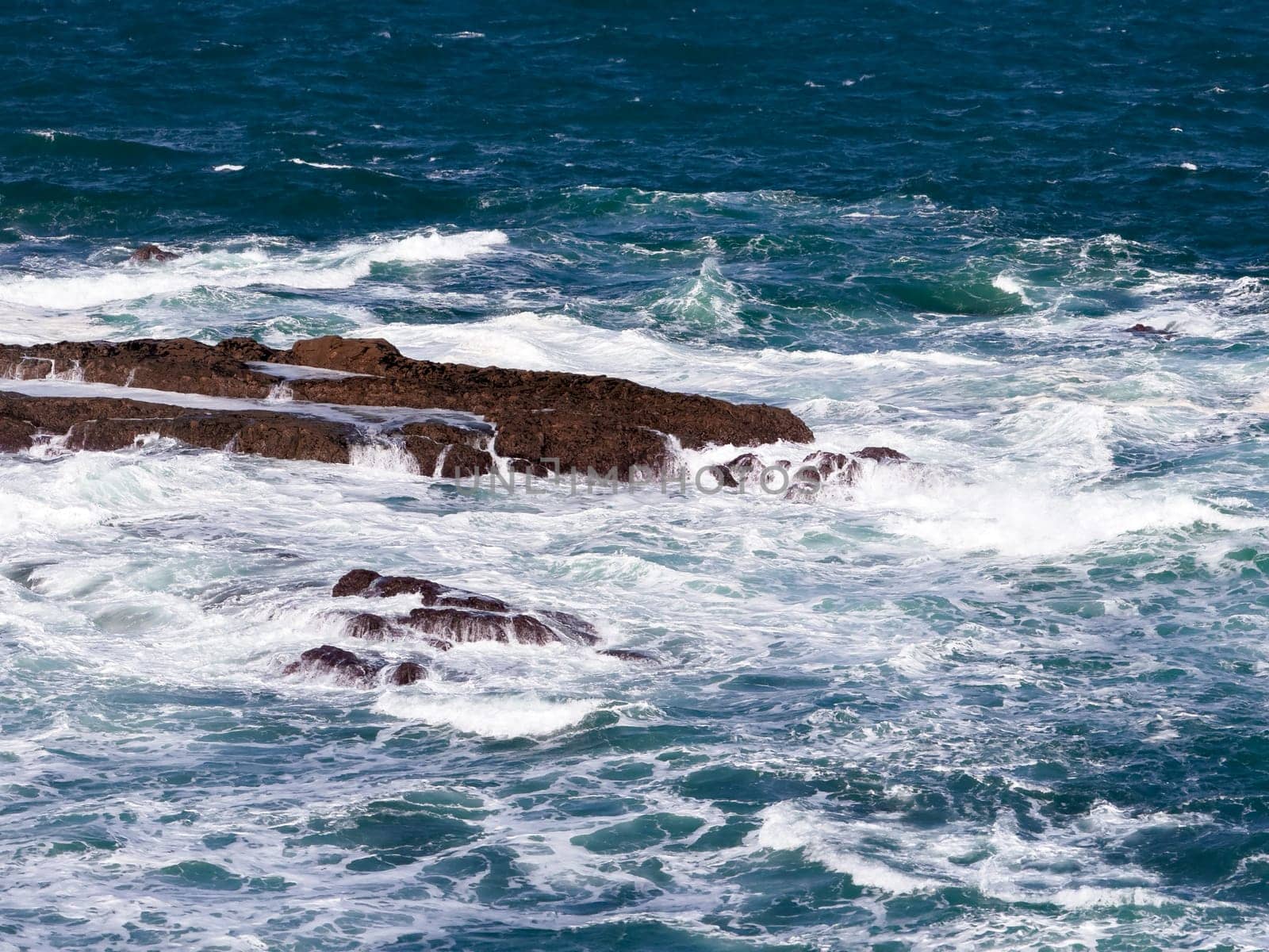 Waves crashing on rocky seashore under clear blue sky.