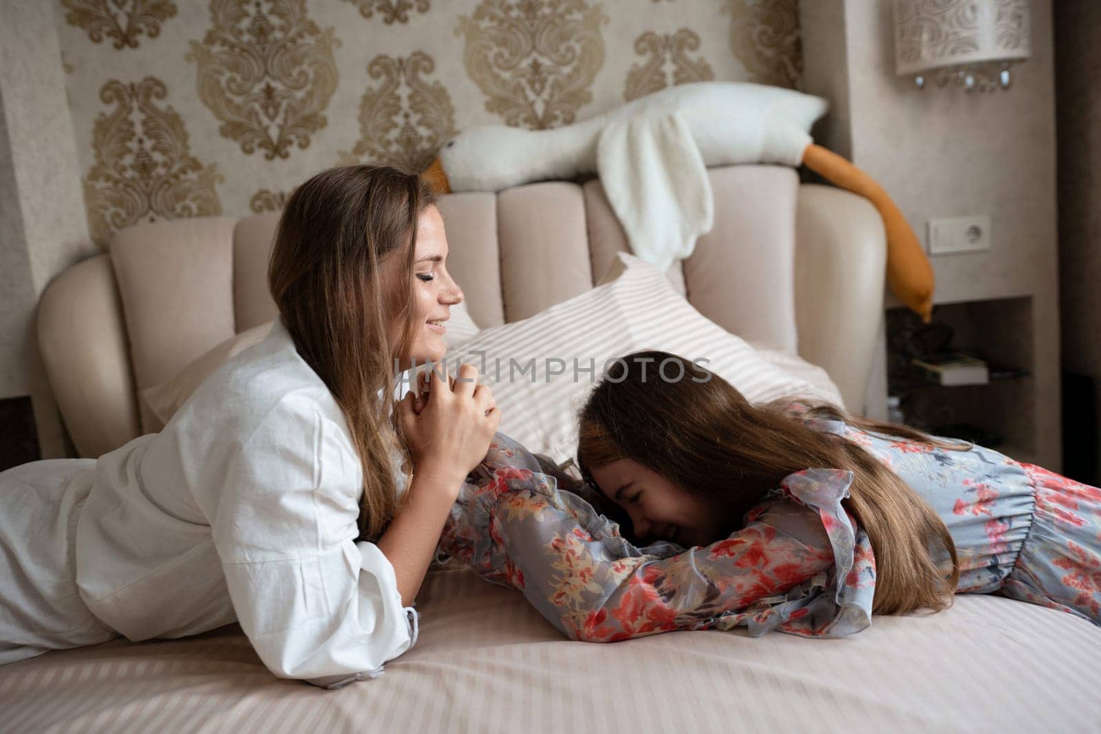 Two women, one in a white robe and one in a patterned dress, lie on a bed. They're close, one leaning toward the other affectionately. Intimate moment of shared closeness.