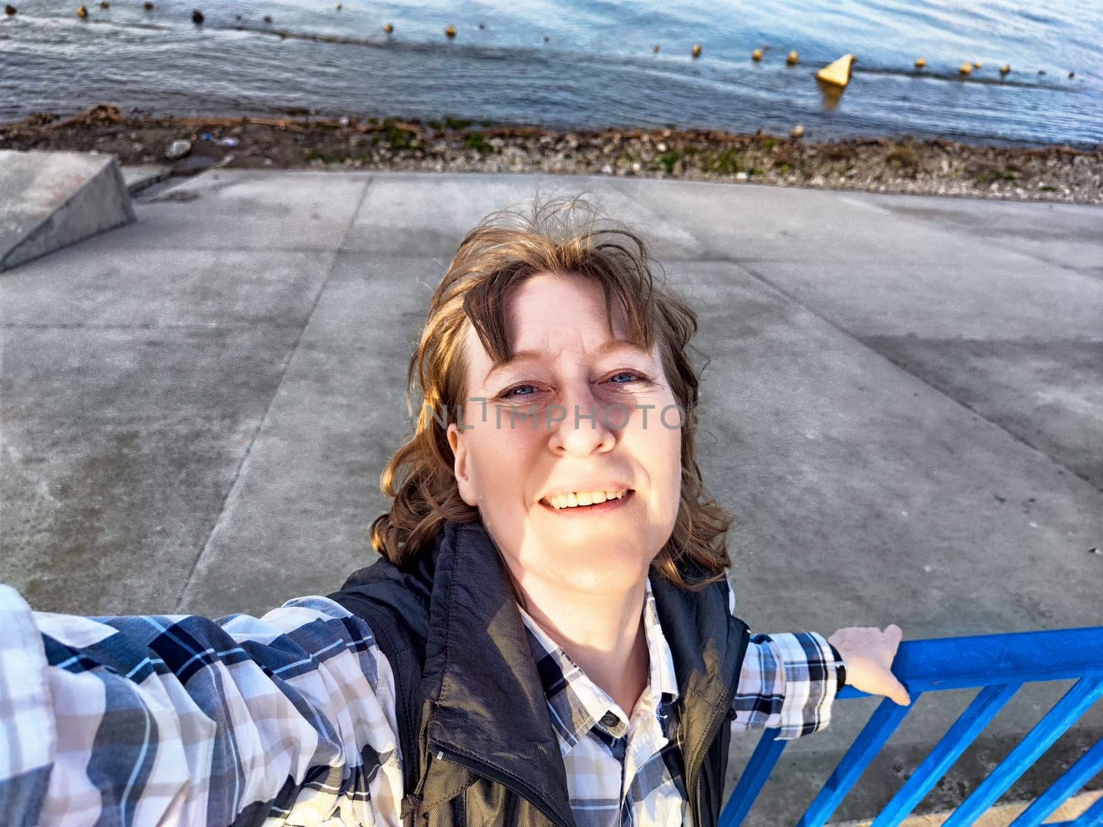 A middle-aged woman enjoying her travel excursion takes a cheerful selfie by the water's edge, embracing nature.