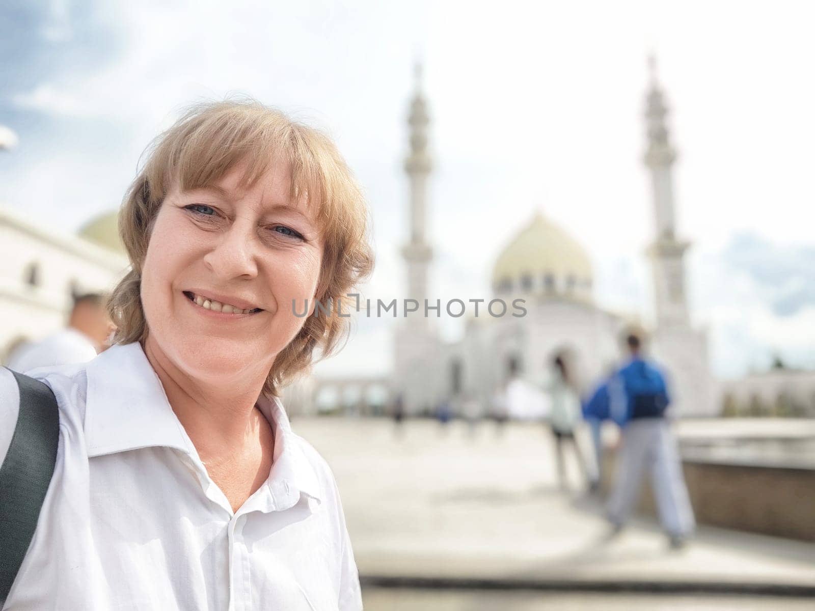 A middle-aged blonde woman smiles for a selfie while exploring a stunning mosque during her travels.
