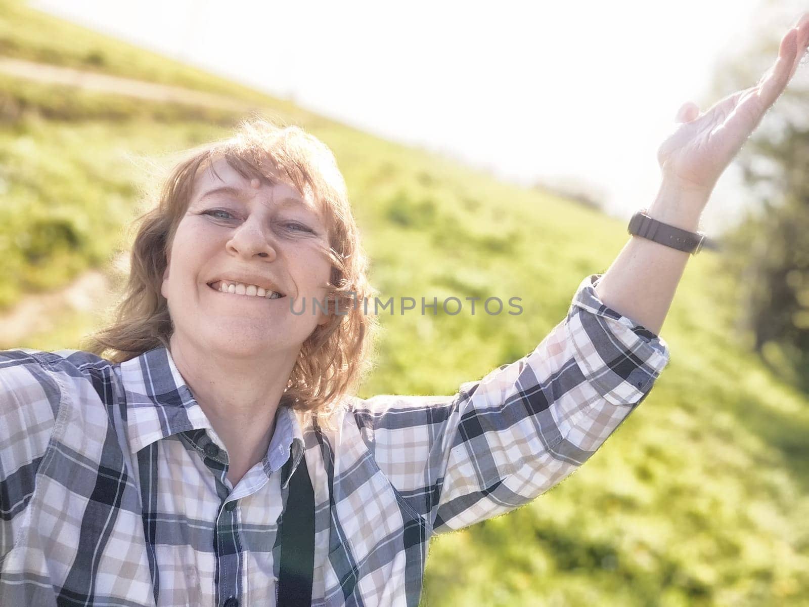 A middle-aged woman with blonde hair smiles brightly as she captures a joyful selfie in a green landscape.