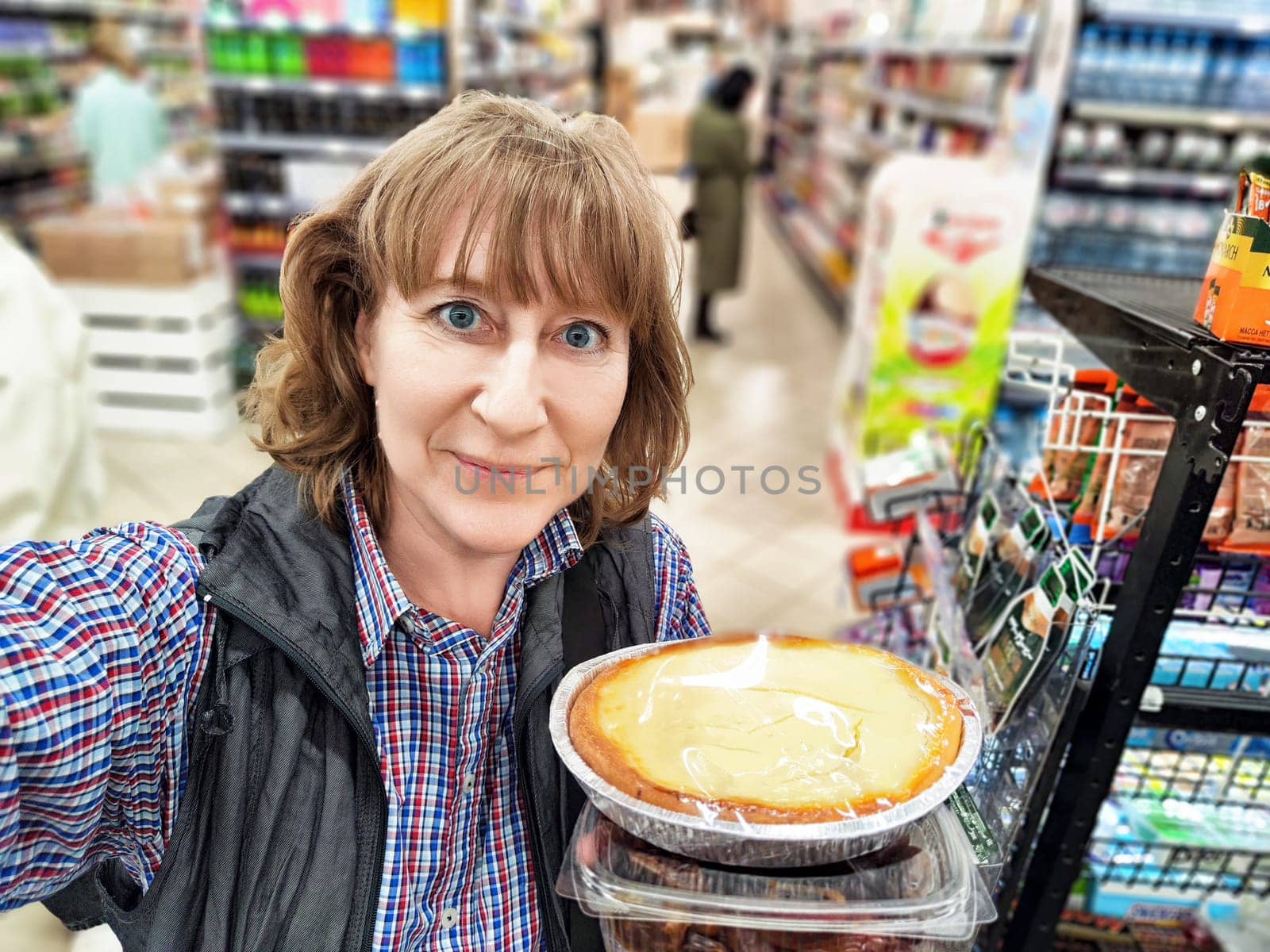 A middle-aged woman captures her shopping trip in a selfie, holding delicious desserts in a lively store environment.