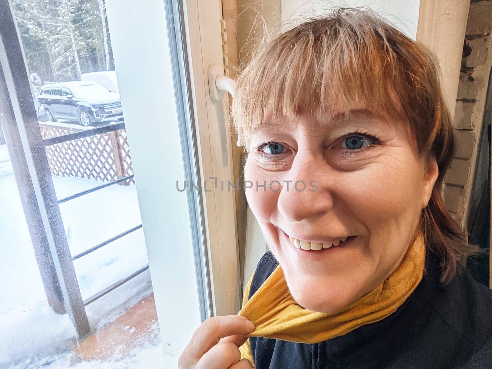 A middle-aged blonde woman captures a cheerful selfie indoors, surrounded by a snowy landscape and trees.