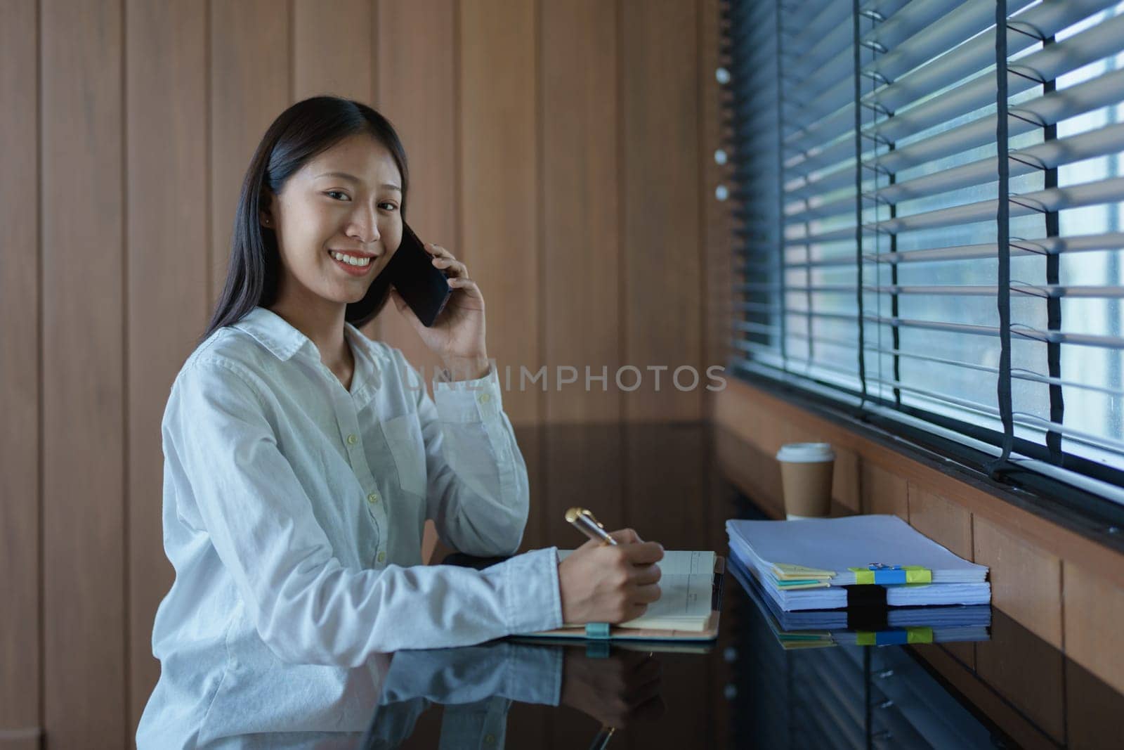 Young woman in casual white shirt smiling while talking on smartphone, working at desk with notepad and coffee.