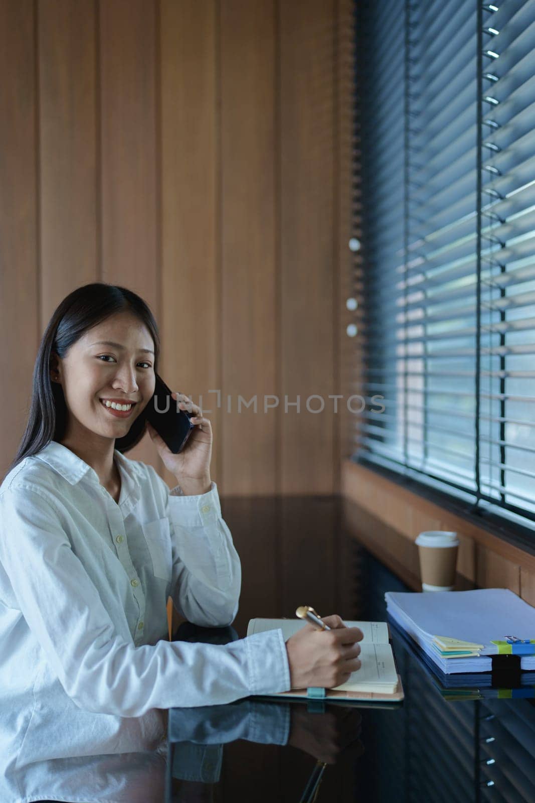 Smiling young woman engaged in a phone conversation while taking notes in a bright, modern office setting.