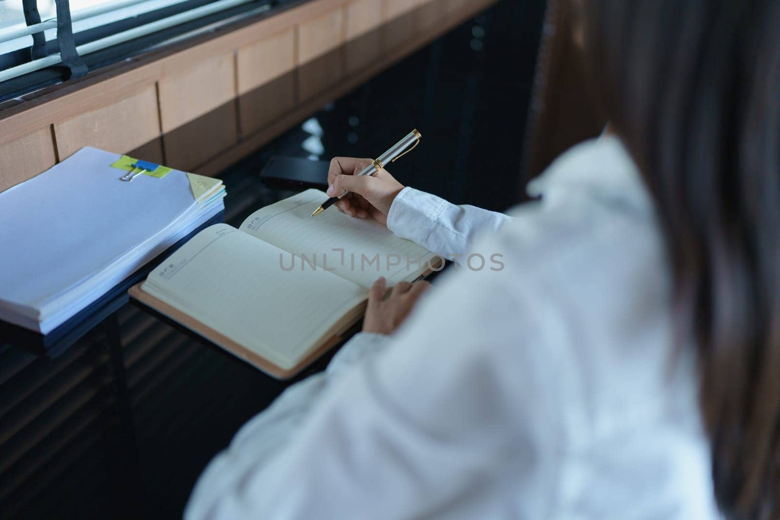 Woman writing in a notebook at a desk with papers and natural light, fostering creativity and productivity.