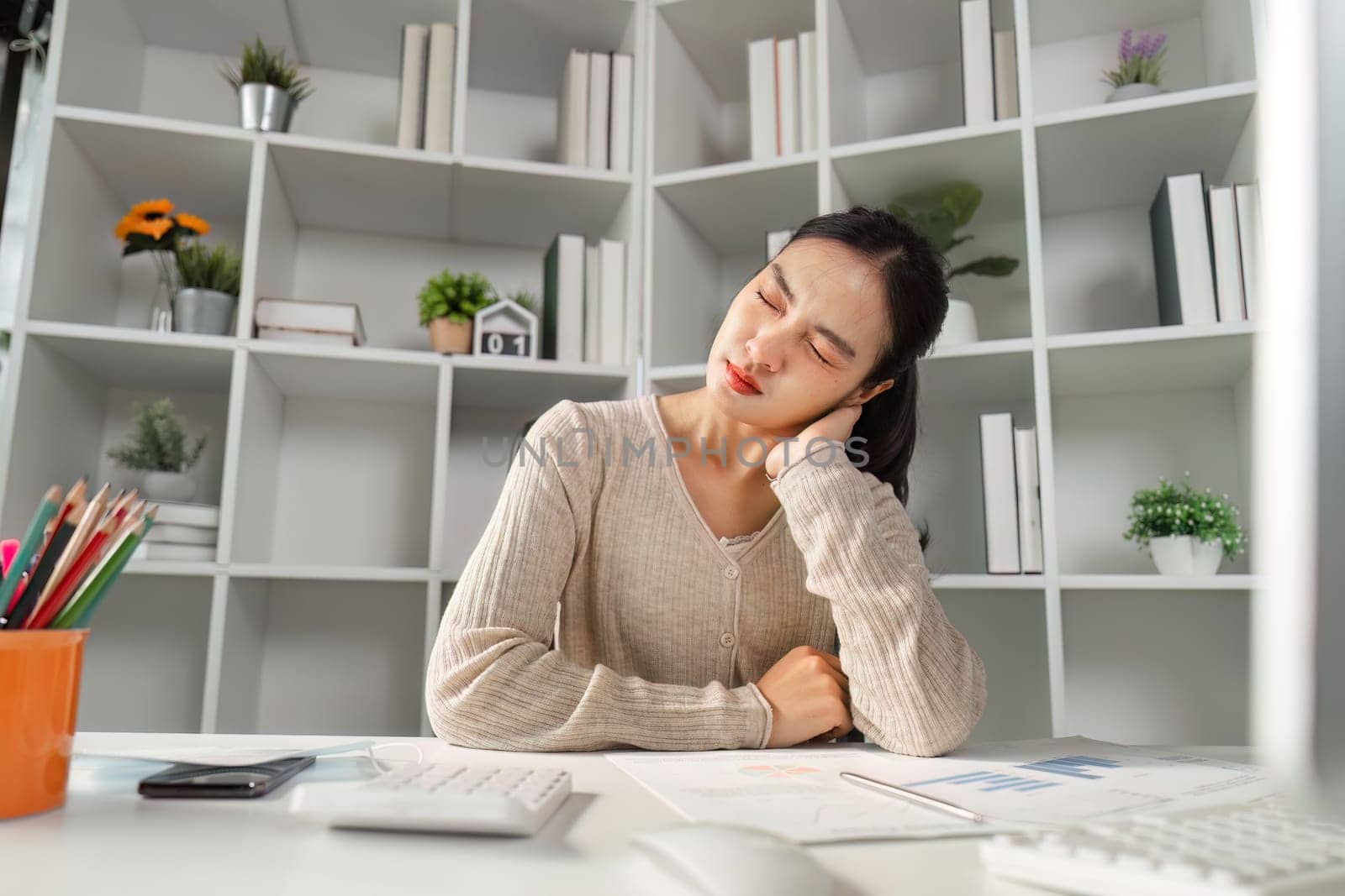 A young woman in a cozy sweater sits at her desk, rubbing her neck in discomfort, surrounded by office supplies and documents.