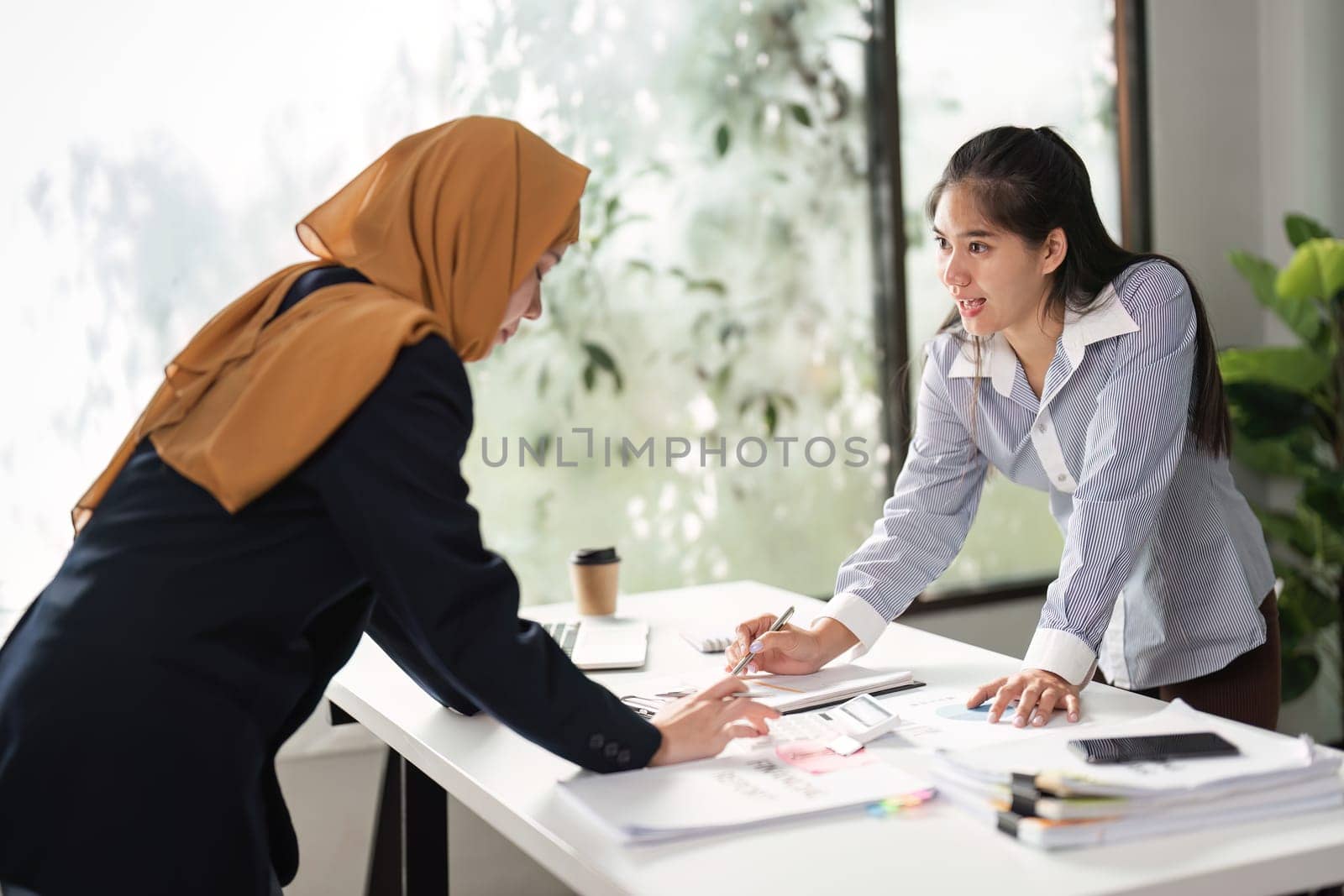 Two women collaborate on financial analysis, discussing reports and strategies in a bright office environment.