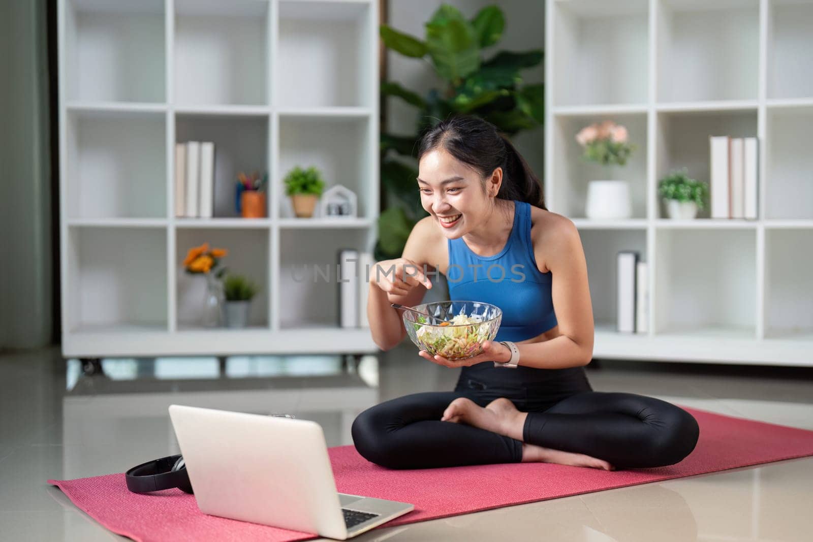 A smiling woman interacts with her laptop while eating a salad, showcasing a blend of technology and healthy eating habits in her living space.