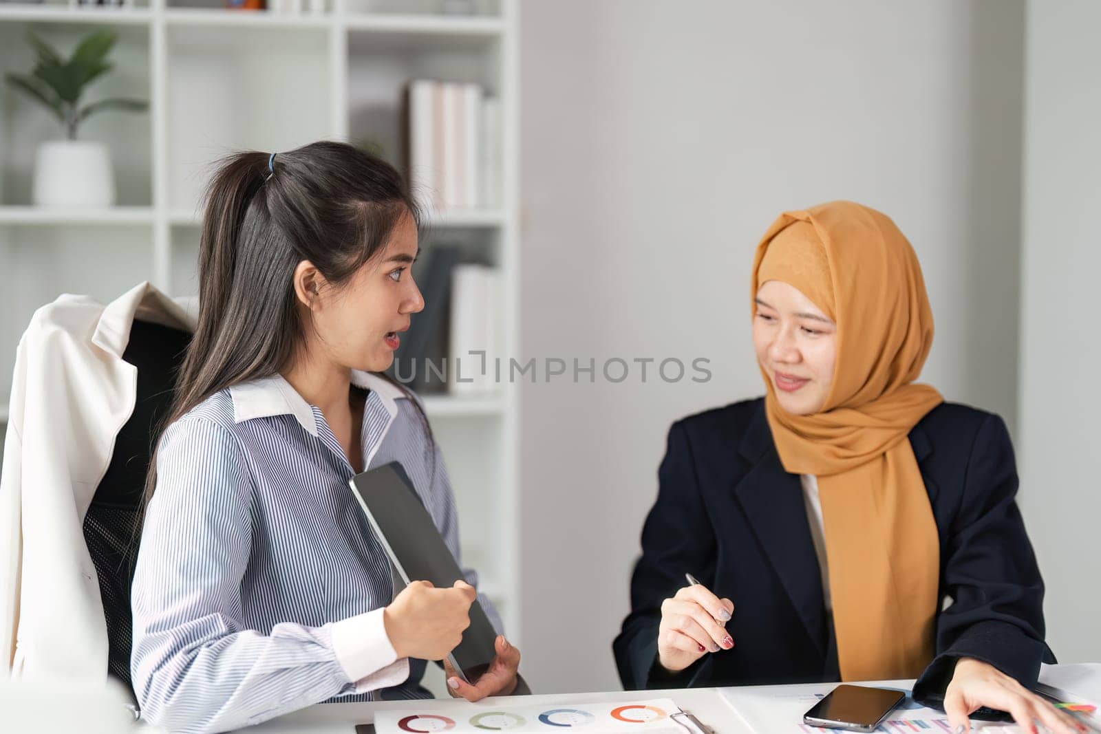 Two diverse women in finance engage in a lively discussion about financial strategies in a contemporary office environment.