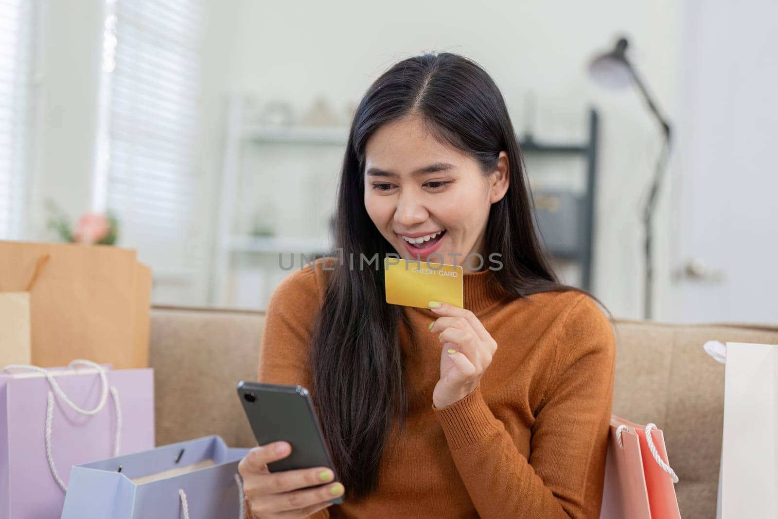 An enthusiastic woman engages in online shopping using her smartphone and credit card, surrounded by colorful shopping bags.