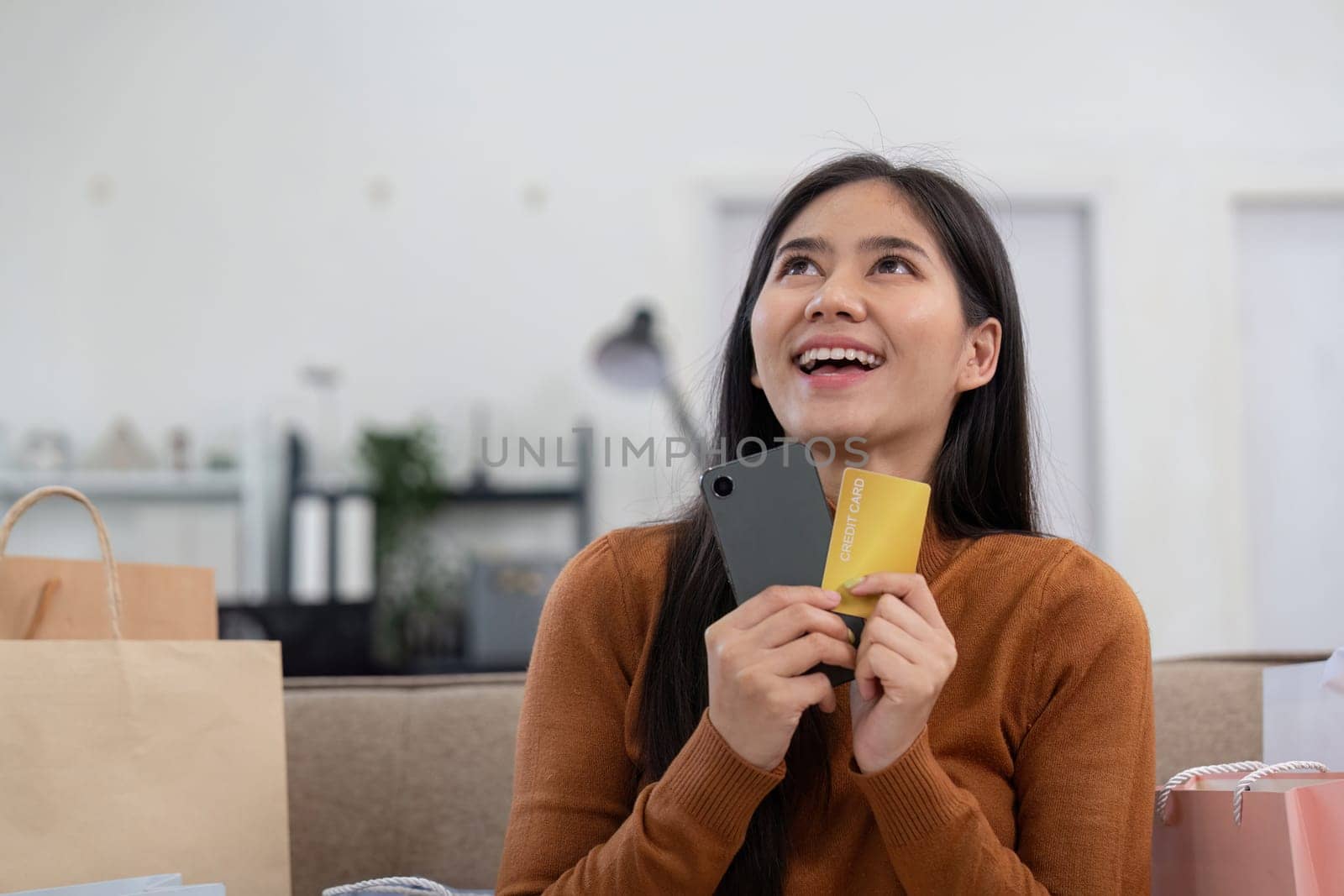 A cheerful woman holds two credit cards and a smartphone, contemplating her online shopping decisions at home.