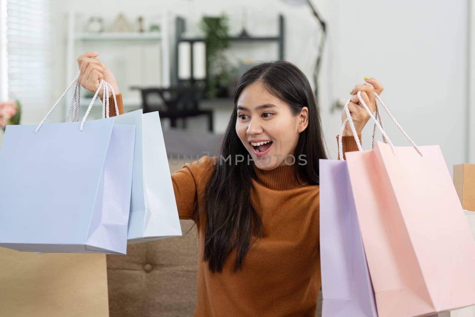 A joyful young woman holds colorful shopping bags while smiling at her smartphone, celebrating her online shopping experience at home.