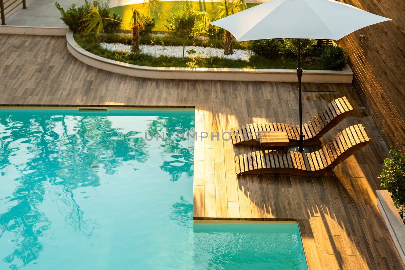 Elegant poolside wooden deck with curved lounge chairs, umbrella, and turquoise pool in a tranquil backyard