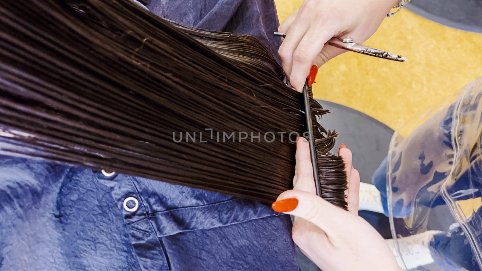 In the bustling atmosphere of a salon, a little girl sits patiently as a stylist with polished red nails carefully trims her damp hair. The sheen of the scissors and the precise hand movements capture the delicate process of styling a child's hair.