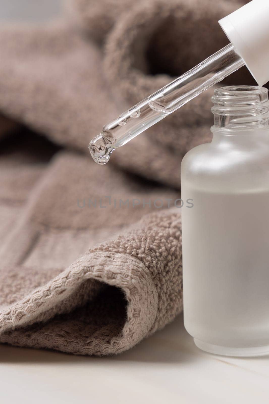 Drop falling from pipette set on opened clear glass dropper bottle near brown bath towel close up. Natural cosmetic serum or essence for facecare, product for Spa and Wellness
