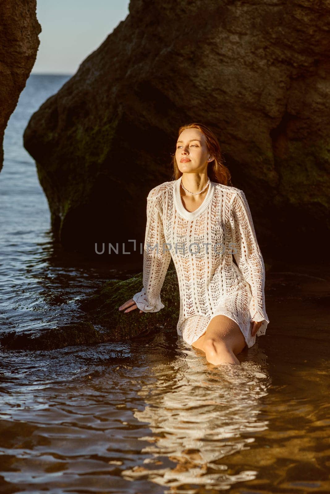 Beautiful young woman posing in the sea, on the beach in summer. girl in the sea on the beach in a white knitted dress. in the water against the backdrop of a rock