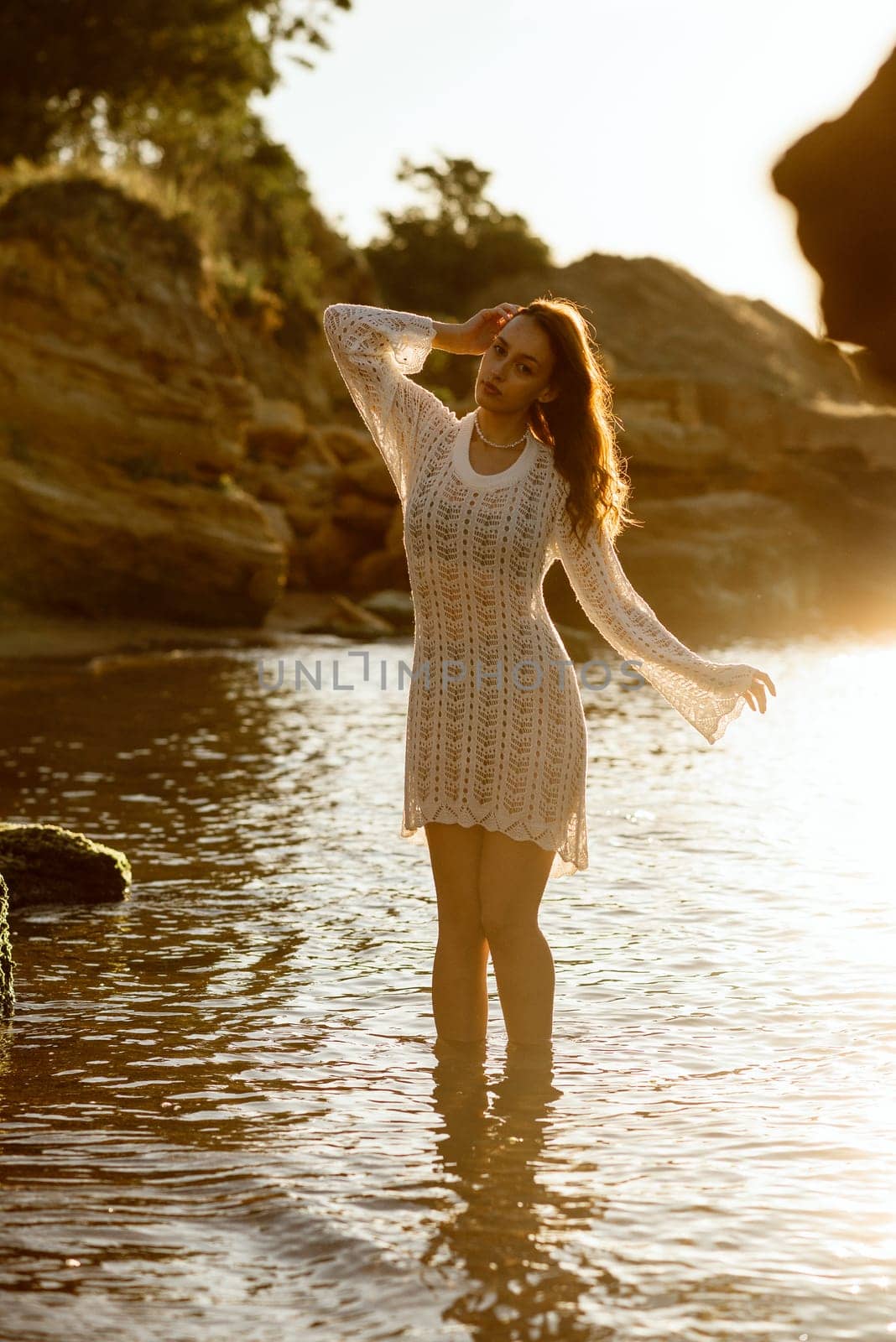 Beautiful young woman posing on the beach in summer. girl standing in backlight. her dress glows in the sun, in white knitted dress with pearls on her neck, in water against the backdrop of a rock