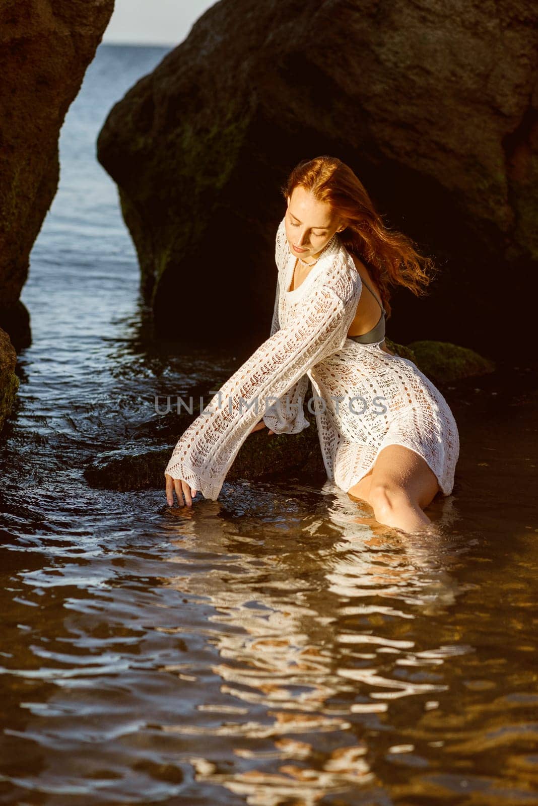 Beautiful young woman posing in the sea, on the beach in summer. girl in the sea sitting on the rocks in a white knitted dress. in the water against the backdrop of a rock