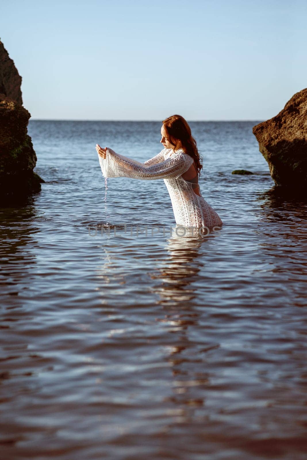 beautiful young girl posing on the sea beach, in the water. she is scooping up sea water with her hands, wearing a white knitted dress, against the backdrop of large rocks. summer sea morning concept